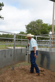 Man in a cowboy hat and plaid shirt standing next to a metal fence with trees in the background