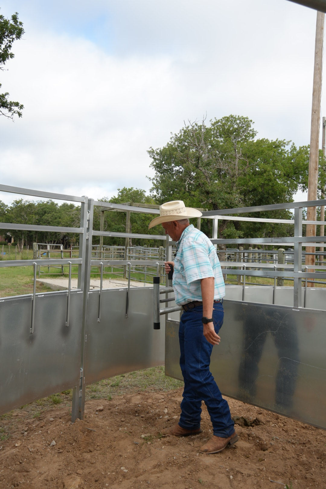 Man in a cowboy hat and plaid shirt standing next to a metal fence with trees in the background